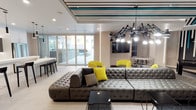 This single-floor amenity space showcases a sophisticated, contemporary design aesthetic defined by bold accents and carefully curated furnishings. The main lounge area is anchored by an oversized tufted gray leather sectional sofa, accented with vibrant yellow and neutral throw pillows. Above, a dramatic multi-arm chandelier serves as a striking focal point, complemented by a striped teal, black, and white ceiling detail that adds visual depth and energy to the space. Yellow accent chairs and small black side tables are thoughtfully arranged throughout, creating distinct seating zones. Large floor-to-ceiling glass doors connect the lounge to an outdoor area, flooding the interior with natural light. A dedicated kitchen area features a futuristic, all-white design with sculptural curved cabinetry, expansive countertops, an integrated sink, stainless steel refrigerator, built-in television, and a cluster of sleek black pendant lights overhead. Dark barstools complete the space. The game room offers a full-size billiard table set against a dramatic dark accent wall adorned with teal and white vertical light installations and a bold geometric ceiling detail. Neutral large-format tile flooring and textured wall art contribute to the polished, upscale ambiance throughout.