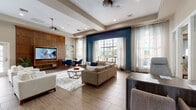 This well-appointed single-floor community space combines comfort and sophistication through a cohesive mid-century modern design aesthetic. The main lounge area is generously proportioned, featuring tray ceilings, recessed lighting, and ceiling fans that contribute to an airy, open ambiance. Wood-look tile flooring runs throughout, complemented by a warm neutral color palette accented by bold navy blue walls that frame oversized windows. These large windows flood the space with abundant natural light and offer views of an outdoor pool area, creating a seamless indoor-outdoor connection dressed with flowing sheer curtains.

The lounge is furnished with multiple comfortable seating arrangements, including plush sofas, armchairs, and a dedicated dining area with navy upholstered chairs. A striking wood-paneled accent wall serves as the backdrop for a large flat-screen television, anchoring the entertainment area. French doors provide additional access to the outdoor areas.

A dedicated business center offers a long built-in desk with multiple computer workstations, illuminated by a statement geometric chandelier overhead, alongside a central round table for collaborative work or meetings. Adjacent to the main lounge, a kitchenette bar features tiled backsplash detailing, dark cabinetry, and bar seating — ideal for casual gatherings. Together, these distinct zones create a versatile, inviting community space designed for both relaxation and productivity.