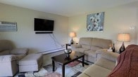 This single-floor space features a thoughtfully arranged living room that serves as its centerpiece. The room is finished with neutral, light-toned walls that create a calm and welcoming ambiance. A wall-mounted flat-screen television with a soundbar serves as a focal point, complemented by a sectional sofa and chaise in a warm beige fabric that provides ample seating. A dark wood coffee table anchors the seating area atop a patterned area rug with earthy tones, adding texture and visual interest to the space. A matching end table holds a decorative lamp that contributes soft, ambient lighting to the room. Wall décor includes a framed floral artwork in muted blue and white tones, adding a subtle artistic touch. A baseboard heating unit runs along the wall, indicating an efficient heating system throughout the space. The overall layout is practical and functional, making efficient use of the available floor area while maintaining an open and comfortable feel. The combination of neutral tones, soft furnishings, and tasteful décor gives the space a relaxed, livable character suited for everyday use.