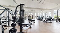 This large-scale fitness center offers a comprehensive workout environment across a single, generously proportioned floor. The space is organized into distinct zones, each designed to accommodate a specific type of training. The main strength area features a robust selection of free weights, cable machines, multi-functional training systems, and adjustable benches, complemented by full-length mirrors and large ceiling fans that enhance airflow and visibility throughout. A dedicated cardio section houses treadmills, air bikes, stair climbers, and stationary cycles positioned in front of tall, narrow windows that frame outdoor views of surrounding greenery, keeping the area bright and motivating. A separate functional training zone provides open floor space for dynamic movement, with medicine balls, plyo boxes, a heavy bag, and additional cardio equipment. Throughout the facility, high ceilings, recessed lighting, and abundant black-framed windows allow natural light to flood the interior, creating a clean, modern aesthetic. Polished concrete-style flooring runs consistently across all zones, offering durability and a contemporary look. A wall-mounted television and built-in storage cabinetry add practical convenience to the space.