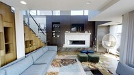This stylish living space showcases a thoughtfully designed interior that blends modern aesthetics with warm, inviting elements. The focal point of the room is a striking dark wood accent wall, which houses a sleek, stone-surround fireplace and a wall-mounted television, creating a cohesive entertainment and comfort hub. Built-in shelving flanks the feature wall, offering both display and storage opportunities.

Natural light is a defining characteristic of this space, with tall clerestory windows positioned high along the upper perimeter, drawing in abundant daylight and creating an airy, open atmosphere. The ceiling height adds a sense of volume and grandeur to the room.

A staircase with glass panel railings and warm wood treads is visible to one side, contributing an architectural detail that reinforces the contemporary design language. The flooring features wide-plank hardwood, adding warmth underfoot.

The living area is furnished with a plush light blue sofa, complemented by a patterned area rug and a mix of green and navy ottomans, introducing color and texture. The overall ambiance is modern, sophisticated, and welcoming.