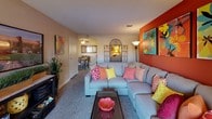 This single-floor residence makes an immediate impression through its bold use of color and energetic design choices. The living room serves as the centerpiece of the space, anchored by a large sectional sofa in a soft grey tone, which provides a neutral contrast to the vivid surroundings. A striking burnt-orange accent wall draws the eye and sets a warm, lively tone, complemented by colorful throw pillows in shades of pink, yellow, and coral.

Decorative wall-mounted ironwork and vibrant artwork further enhance the visual interest of the living area, creating a layered, curated aesthetic. A dark wood coffee table sits atop carpeted flooring, adding grounding contrast to the brighter elements throughout the room.

The layout flows openly toward the dining area, visible through the passage and featuring a ceiling fan, wood dining table, and chairs — connecting the two spaces in a practical and cohesive manner. A textured ceiling and standard-height walls define the interior architecture.

Overall, the space is characterized by its cheerful, energetic palette, comfortable furnishings, and efficient use of an open, connected floor plan.