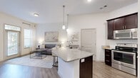 Located at 323 Seven Springs Way in Brentwood, Nashville, TN, this thoughtfully designed one-bedroom, one-bathroom residence offers a comfortable and functional single-floor layout. The open-concept kitchen and living area forms the heart of the home, featuring rich dark-stained cabinetry, granite countertops, a stainless steel refrigerator, and a dishwasher. A central kitchen island with elegant pendant lighting provides additional prep space and casual seating. Wood-style plank flooring runs throughout the main living areas, lending warmth and cohesion to the space. The living room is bathed in natural light from a large door with transom window and offers a relaxed, airy ambiance with generous wall space for décor. The bedroom is carpeted and features vaulted ceilings, a ceiling fan, a large window with sheer curtains that invite natural light, and direct access to both a walk-in closet and the full bathroom. The bathroom includes granite countertops and a shower-tub combination. A dedicated laundry area with built-in shelving accommodates a full-size washer. Clean white walls and a neutral palette throughout create a bright, modern atmosphere suited for everyday living.