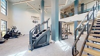 Located at 34 Ramsey Branch Rd, this fitness center is a thoughtfully designed exercise space that combines modern aesthetics with a full suite of workout amenities. The room is defined by its impressive double-height ceilings and expansive grid-pane windows that flood the space with natural light while offering views of the surrounding outdoors. Sleek blue-gray structural columns anchor the open floor plan, while linear LED lighting fixtures overhead contribute to a clean, contemporary ambiance.

The exercise floor is carpeted throughout and generously proportioned to accommodate a wide variety of equipment. Cardio offerings include treadmills, elliptical machines, a Spirit stair climber, and a stationary bike. Strength training is well represented with cable machines, a multi-functional weight rack, a Smith machine, benches with free weights, and additional resistance equipment. A dedicated area near one wall stores medicine balls, resistance bands, and dumbbells, supporting functional and free-weight training.

A staircase with a modern metal railing leads to an upper mezzanine level, visible from the main floor and fitted with additional equipment, maximizing the usable training area. Motivational wall graphics, including the phrase "Stronger Than Yesterday," add an energizing character to the space, making this fitness center as inspiring as it is functional.