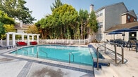 Located at 1235 Ashbourne Rd in Elkins Park, Pennsylvania, this two-floor property features a well-appointed outdoor pool area that serves as a central amenity of the community. The generously sized swimming pool is finished with classic blue tile accents along its edges and is equipped with stainless steel entry railings, providing both style and accessibility. Surrounding the pool is a spacious poured concrete and paver sundeck outfitted with multiple lounge chairs and chaise lounges, ideal for relaxing in the sun. A white pergola structure anchors one corner of the deck and is paired with vibrant red outdoor sofas, creating a shaded lounge area with a pop of color. Additional outdoor dining and seating areas with umbrella tables are positioned nearby, offering options for al fresco gatherings. The entire pool area is enclosed by a crisp white picket fence, lending a clean and classic aesthetic to the space. Towering mature trees frame the perimeter, providing natural privacy screening and greenery throughout the seasons. The multi-story residential buildings with traditional siding and chimney details are visible in the background, giving context to the broader community setting. The overall ambiance is bright, open, and inviting, with a resort-like quality enhanced by the lush natural surroundings.