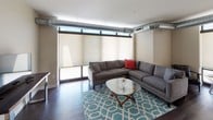 This single-floor residence offers a thoughtfully designed open-concept layout that seamlessly connects the kitchen, dining, and living areas. The kitchen is a standout feature, showcasing light maple cabinetry, dark granite countertops, a center island with pendant lighting overhead, and a full suite of stainless steel appliances including a refrigerator, gas range, built-in microwave, and dishwasher. Rich dark hardwood flooring runs throughout the main living areas, adding warmth and contrast to the neutral color palette.

The living room is anchored by large clerestory windows that allow natural light to filter in, complemented by exposed industrial-style ductwork that lends a modern, urban character to the space. A sliding door provides access to an outdoor area, further enhancing the connection between interior and exterior.

The bedroom is carpeted for comfort and features high-positioned windows that offer privacy while still admitting natural light. A wall-mounted air conditioning unit ensures year-round climate control. The bathroom is clean and functional, with a wood-toned vanity, white countertop, a tub-shower combination, and contemporary fixtures. The entryway features the same dark hardwood flooring and includes a thermostat panel, offering a welcoming and practical transition into the home.