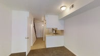 This two-story residence offers a straightforward and functional layout designed for practical everyday living. The main level features an open-concept living and kitchen area, where the two spaces flow seamlessly into one another. The kitchen is equipped with a white electric range, a sink set into a light-toned countertop, and white cabinetry above, creating a clean and cohesive look. A peninsula-style counter defines the boundary between the kitchen and living area, adding a sense of structure to the open floor plan. The living area is finished with wall-to-wall carpeting, providing a warm and comfortable feel underfoot, while the kitchen and hallway transition to tile flooring for easy maintenance. A textured popcorn ceiling runs throughout the main level, and flush-mounted ceiling lighting provides consistent illumination. A hallway connects the main living space to the front entryway, where a classic paneled door with brass hardware welcomes residents and guests. A staircase at the end of the hallway leads to the upper floor, expanding the overall living capacity of the home. The HVAC vent integrated into the ceiling ensures year-round climate comfort throughout the space.