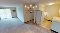 This single-floor residence offers a straightforward, functional layout designed for comfortable everyday living. The open-concept living and dining area is finished with neutral wall-to-wall carpeting that lends warmth to the space, while clean, light-colored walls create a bright and airy atmosphere throughout. A classic candelabra-style chandelier serves as a focal point, anchoring the dining area with a touch of traditional elegance. The kitchen is positioned adjacent to the living and dining space, offering easy accessibility and a natural flow between areas. It features white cabinetry, a full-size refrigerator, and a white electric range, all set on light-toned flooring that contrasts neatly with the carpeted living areas. The kitchen layout is efficient and well-organized, providing ample storage and workspace. A wall-mounted air conditioning unit is visible in the living area, ensuring year-round comfort. The overall design is clean and neutral, offering a versatile canvas well-suited to a variety of furnishing styles. The space is practical and move-in ready, with each area defined clearly within the open floor plan.