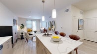 This well-appointed single-floor residence showcases a bright, open-concept design with a seamless flow between the kitchen, dining, and living areas. The kitchen is a standout feature, boasting an expansive quartz island with striking veining, light wood cabinetry extending to the ceiling, white quartz countertops, a full suite of stainless steel appliances, and a deep undermount sink. Stylish pendant lighting illuminates both the island and the dining area, enhancing the modern aesthetic.

The open living and dining space is bathed in natural light from large windows with views to the outdoors. Light wood-style flooring runs throughout, complementing the neutral color palette. The living area offers a comfortable, contemporary atmosphere anchored by a curved sofa and vibrant abstract artwork.

The bedroom provides a serene retreat with ample natural light from well-dressed windows, a ceiling fan, and generous wall space for artwork and storage. The bathroom features a dual-sink vanity with light wood cabinetry, white quartz countertops, a large wall mirror, and decorative tile accents, offering both functionality and refined style. Together, these spaces create a cohesive, modern living environment.