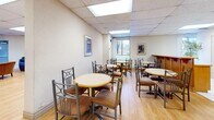 This single-floor space offers a thoughtfully organized layout designed to support both social gatherings and quiet relaxation. The main dining area is a welcoming, open room featuring light hardwood flooring, a suspended acoustic tile ceiling with recessed panel lighting, and multiple round tables with metal-frame chairs arranged to encourage communal dining. Framed artwork decorates the neutral-toned walls, and windows allow natural light and outdoor views of surrounding trees and greenery to filter in.

The kitchen is fully equipped with warm-toned wood cabinetry, a white tile backsplash, light tile flooring, and a full suite of appliances including a refrigerator, electric stove, microwave, and double sink — providing ample functionality for food preparation and service.

Adjacent lounge areas offer comfortable seating arrangements, including upholstered wingback chairs with decorative side tables and a separate sitting area with leather sofas and armchairs. These spaces are accented with artwork, area rugs, and natural light from casement windows. Together, the zones create a versatile, community-oriented environment that balances practical amenities with a warm, welcoming ambiance throughout.