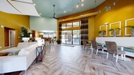 Located at 6465 142nd Avenue North in Clearwater, Florida, this single-floor community clubhouse offers a generously sized, thoughtfully designed gathering space ideal for both social and professional use. The interior is defined by a bold color palette of warm golden-yellow walls contrasted with soft teal ceilings, creating an energetic yet cohesive aesthetic throughout.

The open-concept main area seamlessly connects the living room, dining space, and kitchen. The living room features soaring vaulted ceilings, a wall-mounted flat-screen TV, a plush white sectional sofa, and eclectic accent seating. Herringbone-patterned wood-look tile flooring flows throughout, adding a sophisticated touch. Large sliding glass doors flood the space with natural light and provide access to an outdoor covered lounge area, which is equipped with high-top seating, a vibrant sectional sofa, and multiple ceiling fans — all surrounded by rows of tall windows overlooking the outdoors.

The kitchen includes a large central island with bar seating, pendant lighting, white cabinetry, dark countertops, and a vivid teal mosaic tile backsplash. Adjacent dining areas feature built-in banquette seating with decorative wall framing.

The dedicated office provides a professional conference-style setting with a glass table, ergonomic white chairs, built-in workstations with computers, and tasteful artwork — making it a fully functional workspace within the clubhouse.