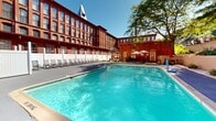 Located at 155 W Main St in Vernon Rockville, Connecticut, this outdoor space is defined by the dramatic presence of a historic red brick mill building that serves as its architectural backdrop. The multi-story structure features rows of large arched windows and a distinctive steeple, reflecting the region's industrial heritage and lending the area a sense of grandeur and character.

The centerpiece of the outdoor area is a generously sized swimming pool with clear blue water, framed by a spacious paved pool deck. A long row of lounge chairs lines one side of the pool, offering ample space for relaxation in the open air. A decorative fountain feature rises from the pool, adding a lively visual element to the setting. Additional seating areas with umbrella-shaded tables and chairs are positioned throughout the deck, providing comfortable spots for outdoor dining or leisure.

The space is enclosed by white privacy fencing, creating a defined and inviting atmosphere while the surrounding mature trees provide natural shade and a verdant backdrop. The overall ambiance blends historic industrial architecture with a well-appointed recreational outdoor environment, making it a distinctive and functional amenity suited for warm-weather enjoyment.
