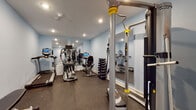 This dedicated fitness space is thoughtfully designed to support a full range of exercise routines. The room is finished with light blue walls and recessed lighting overhead, creating a clean, energizing atmosphere. Durable rubber flooring lines the entire space, providing both safety and comfort during workouts.

The cardio section is outfitted with commercial-grade True Fitness equipment, including a treadmill, elliptical trainer, upright stationary bike, and a recumbent bike — each featuring an interactive display console for performance tracking. A large wall-mounted mirror spans one end of the room, visually expanding the space and allowing users to monitor their form.

Strength training is equally well-served. A multi-functional cable training system with adjustable attachments anchors one side of the room, while a tiered dumbbell rack stocked with a full range of free weights occupies an adjacent corner. An adjustable workout bench and a step platform round out the strength and conditioning equipment.

Additional amenities include a wall-mounted functional training reference chart, a small storage shelf, and direct access via two doors — one of which features a window that allows natural light to filter into the space. The overall layout is organized and efficient, making this fitness room a capable and professional training environment.