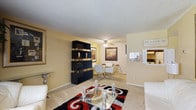 This single-floor space presents an open-plan layout that connects the living and dining areas in a cohesive, functional arrangement. The living area is anchored by a bold red, black, and white patterned area rug beneath a glass coffee table, adding a striking visual contrast to the light-toned carpet flooring that extends throughout. A standout feature is the freestanding black and gold shelving unit, which serves as a stylish room divider, elegantly separating the living space from the dining area without fully enclosing either zone. The dining area accommodates a white table with modern chairs and is set against a textured, subtly patterned accent wall adorned with framed artwork, adding warmth and character. A ceiling fan overhead provides both practical ventilation and a finishing touch to the space. A pass-through window connects to an adjacent kitchen area, enhancing the open feel and allowing for easy interaction between spaces. The textured ceiling and warm yellow-toned walls give the interior a cozy atmosphere throughout, while wall art and decorative accents contribute to an intentional, styled aesthetic.