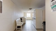 This shared laundry facility is designed with functionality and convenience in mind. The single-floor space features a clean, open layout with light-toned tile flooring that runs the full length of the room, making it easy to navigate and maintain. Along one side, a row of top-load washing machines is neatly arranged, while the opposite side houses a series of stacked washer and dryer units, maximizing the available floor space and providing ample laundry capacity. A white wooden bench is positioned along the wall, offering a comfortable place for residents to wait while their laundry cycles run. The room is illuminated by overhead lighting, and a window at the far end allows natural light to filter in, contributing to a bright and airy atmosphere. The walls feature a textured finish in a neutral tone, and ventilation is provided through ceiling vents, keeping the space well-aired. The overall ambiance is practical and tidy, with a straightforward design focused on meeting the everyday laundry needs of its users efficiently.