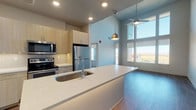 This single-floor unit showcases a contemporary open-concept design with notable architectural variety. The kitchen serves as a central feature, equipped with stainless steel appliances including a refrigerator, range, and built-in microwave. Light wood cabinetry extends floor-to-ceiling, complemented by white quartz countertops and a large waterfall island with an integrated sink. A stylish pendant light anchors the space above the island. The living area adjacent to the kitchen benefits from dramatically tall ceilings and an expansive window wall that floods the space with natural light and frames sweeping panoramic views of the surrounding landscape. A ceiling fan provides additional comfort.

The bedrooms are carpeted and well-proportioned, each featuring large windows that capture outdoor views. One bedroom includes built-in wood shelving and cabinetry for added storage. Two bathrooms serve the unit: one features a tiled soaking tub and shower combination, while the other offers a glass-enclosed walk-in shower. Both include light wood vanities with quartz countertops and wood-look tile flooring. A dedicated laundry room with a full-size washer and dryer, overhead shelving, and a built-in cabinet rounds out this well-appointed space.