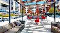 Located at 200 Pleasant St in Malden, Massachusetts, this single-level outdoor amenity space offers a thoughtfully designed environment for relaxation and recreation. The space is framed by a contemporary building facade featuring a striking combination of deep red and neutral paneling with large windows, lending an urban, modern character to the setting.

The outdoor area is organized into several distinct zones, each designed for a different type of leisure. A hammock lounge area provides a casual retreat, with multiple blue rope hammocks suspended between freestanding frames on a clean tiled surface, surrounded by mature trees and flowering shrubs. Nearby, a pergola structure supports a series of bold red hanging swing chairs alongside wicker seating, creating a vibrant, social gathering point bathed in natural sunlight.

Throughout the space, warm-toned hardwood decking and horizontal wood-slat privacy screens with dark steel frames define separate areas while allowing light and views to filter through. These screens add a refined architectural detail that balances openness and privacy. Oversized chess sets and additional lounge furniture complete the space, contributing to an amenity-rich outdoor environment suited for both active and relaxed use.