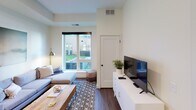 This single-floor residence offers a thoughtfully designed layout that balances comfort and functionality. The kitchen is outfitted with white shaker-style cabinetry, granite countertops with subtle veining, a subway tile backsplash, and hardwood-style flooring, creating a clean and practical workspace. A stainless steel sink and modern hardware complete the polished look, while the entry door adds a warm contrast with its rich dark wood finish. The living area is bright and inviting, featuring hardwood-style flooring, a large window that draws in natural light and frames views of the surrounding exterior, and a neutral color palette. The space comfortably accommodates a sectional sofa, a wood coffee table, and an entertainment console, making it ideal for everyday living. A ceiling fan is present in the bedroom, which is carpeted and features soft, muted wall tones, a large window overlooking a landscaped courtyard area, and ample space for a dresser and seating. The bathroom is clean and accessible, finished in white tile with grab bars, a shower with a curtain, and generous floor space. Throughout the unit, recessed and ceiling-mounted lighting, neutral walls, and consistent finishes create a cohesive and modern ambiance.