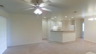 This single-floor residence at The Brickyard offers an open-concept layout that seamlessly connects the living area to the kitchen. The living space features neutral carpet flooring and is topped with a ceiling fan, providing both comfort and airflow. Recessed lighting is installed throughout, ensuring consistent, even illumination across the space. A standard interior door provides access to an adjacent room or hallway, contributing to the functional flow of the floor plan. The kitchen is positioned toward the rear of the space and features a raised breakfast bar countertop that serves as a natural divider between the cooking and living areas. White cabinetry lines the kitchen walls, offering ample storage, while the white refrigerator complements the clean, neutral palette of the space. The kitchen also benefits from its own recessed lighting, maintaining the cohesive lighting design found throughout. The overall color scheme is light and neutral, with white walls and light-toned finishes contributing to a bright, airy ambiance. The open layout and connected design make the space feel practical and easy to navigate.