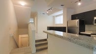 This three-floor space showcases a thoughtfully designed interior that blends functionality with modern finishes. The kitchen is a standout area, featuring granite countertops with a classic speckled pattern, sleek dark lower cabinetry, and stainless steel appliances including a full-size refrigerator. Track lighting mounted on a raised ceiling section illuminates the kitchen, adding a contemporary touch while enhancing visibility across the space.

The open layout creates a natural flow between the kitchen and the surrounding living areas, with light tile flooring extending throughout the main level. Carpeted stairs connect the floors, transitioning between levels with ease. White walls and ceilings throughout contribute to a clean, bright aesthetic, while multiple windows fitted with horizontal blinds allow natural light to filter in.

Architectural details such as the varied ceiling heights and a raised platform area add visual interest and a sense of depth to the layout. The multi-level design provides distinct zones within the space, making it both versatile and well-structured for everyday living across all three floors.