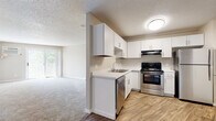 This single-floor residence offers a clean, functional layout designed for comfortable everyday living. The kitchen is a standout feature, outfitted with crisp white shaker-style cabinetry, light countertops, and a full suite of stainless steel appliances including an electric range, dishwasher, and refrigerator. Wood-look flooring runs throughout the kitchen, providing a warm contrast to the bright cabinetry. The kitchen opens directly into the living area, which is carpeted and bathed in natural light streaming through a sliding glass door that leads to a private balcony overlooking lush greenery. Baseboard heating runs along the perimeter of the living space, and a wall-mounted air conditioning unit ensures year-round comfort. The bedroom is generously sized with plush carpet, an open reach-in closet with built-in shelving and hanging rods, and a nearby walk-in storage closet. The full bathroom is finished with classic white tile flooring, a single-sink vanity with white cabinetry, and a tub-shower combination. Additional built-in shelving adjacent to the bathroom provides ample linen and storage space. Textured ceilings and neutral wall tones throughout give the space a cohesive, move-in-ready feel.