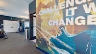 This state-of-the-art fitness center, located at 249 Allure Ln in Chesapeake, VA, spans 1630 square feet on a single floor. The space is thoughtfully divided into distinct workout areas, each tailored to specific fitness needs. The cardio zone boasts a row of treadmills and elliptical machines facing large windows, allowing natural light to flood the area. A separate strength training section features an array of weight machines and free weights, complete with a full-length mirror wall for form checks.

The facility's design incorporates a modern aesthetic with dark blue walls contrasting against white ceilings, creating a sleek and energizing atmosphere. Motivational wall art, including a large mural with the phrase "Doesn't challenge you, doesn't change you," adds inspiration to the space. High ceilings with exposed ductwork and contemporary ceiling fans contribute to the open, airy feel.

Additional amenities include a dedicated stretching area with mats, a functional training space with equipment like battle ropes and medicine balls, and a separate room that could serve as a studio for group classes or personal training. The center also features a small lounge area with comfortable seating and a children's play corner, making it family-friendly. Throughout the space, strategically placed televisions and sound systems enhance the workout experience, while ample lighting ensures visibility and safety during exercise routines.