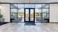This single-story clubhouse is designed with a sophisticated, contemporary aesthetic that balances bold design choices with welcoming functionality. The entry foyer immediately sets the tone, featuring a expansive floor-to-ceiling glass curtain wall with dark steel framing and double glass doors that flood the space with natural light while offering unobstructed views of the outdoor courtyard, where string lights and manicured landscaping are visible. A branded accent rug anchors the tiled entry floor, and flanking bench seating provides a polished first impression.

Moving inward, a glass sliding door leads to a leasing or meeting area distinguished by a striking crystal chandelier, dark veined marble flooring, and a marble-topped reception counter. Adjacent to this, the main lounge area commands attention with soaring vaulted ceilings accented by bold navy exposed beams and an elegant candelabra-style chandelier. Large picture windows and French doors on multiple walls bathe the seating area in natural light and offer views of the surrounding grounds.

The kitchen-style bar area is a design standout, featuring vibrant emerald-green cabinetry, a marble-topped island, mirrored glass cabinet fronts, and dramatic Moroccan-inspired pendant lights. Nearby, a beverage station presents a geometric-patterned tile accent wall, gray cabinetry, and a branded drink dispenser, completing the refined amenity experience throughout this thoughtfully curated space.