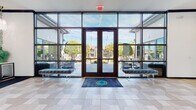 This single-story clubhouse is designed with a sophisticated, contemporary aesthetic that balances bold design choices with welcoming functionality. The entry foyer immediately sets the tone, featuring a expansive floor-to-ceiling glass curtain wall with dark steel framing and double glass doors that flood the space with natural light while offering unobstructed views of the outdoor courtyard, where string lights and manicured landscaping are visible. A branded accent rug anchors the tiled entry floor, and flanking bench seating provides a polished first impression.

Moving inward, a glass sliding door leads to a leasing or meeting area distinguished by a striking crystal chandelier, dark veined marble flooring, and a marble-topped reception counter. Adjacent to this, the main lounge area commands attention with soaring vaulted ceilings accented by bold navy exposed beams and an elegant candelabra-style chandelier. Large picture windows and French doors on multiple walls bathe the seating area in natural light and offer views of the surrounding grounds.

The kitchen-style bar area is a design standout, featuring vibrant emerald-green cabinetry, a marble-topped island, mirrored glass cabinet fronts, and dramatic Moroccan-inspired pendant lights. Nearby, a beverage station presents a geometric-patterned tile accent wall, gray cabinetry, and a branded drink dispenser, completing the refined amenity experience throughout this thoughtfully curated space.