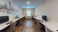 This single-floor business center offers a clean and functional workspace designed to accommodate multiple users simultaneously. The room is laid out with long, sleek white desks running along both walls, each equipped with desktop computers, keyboards, and mice, providing ample workstation space for residents or guests. Comfortable upholstered rolling chairs accompany each station, ensuring an ergonomic and professional setting.

The flooring throughout the space features rich, dark hardwood-style planks that add warmth and contrast against the light-toned walls. Recessed ceiling lighting provides consistent, even illumination, while a window dressed with horizontal blinds allows for natural light and a connection to the outdoors. A cityscape mural decorates one wall, adding a distinctive urban aesthetic that enhances the overall character of the room.

Near the entrance, a printer is conveniently accessible, rounding out the practical amenities of the center. The space is accessed through double glass-panel doors, contributing to an open, transparent feel at the entry point. A posted notice board near the exit provides informational signage for users. The business center is thoughtfully designed to balance function and style, making it an efficient and inviting environment for work tasks.
