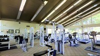 This fitness center offers a comprehensive workout environment housed within a single-story structure characterized by an exposed beam ceiling and abundant natural light. Large windows line the perimeter walls, providing views of the surrounding greenery and creating an airy, open atmosphere that enhances the workout experience.

The facility is generously proportioned, accommodating a wide variety of exercise equipment without feeling cramped. The strength training area features multiple cable and pulley machines, weight benches, and resistance stations, all finished in white, giving the space a clean, organized appearance. The cardio section is equally well-appointed, offering upright and recumbent stationary bikes, treadmills, ellipticals, and rowing-style machines arranged to allow comfortable circulation between equipment.

Full-length mirrors are mounted along the walls, amplifying the sense of space and allowing users to monitor their form during workouts. Flooring consists of a patterned tile or rubber mat combination, providing both durability and safety underfoot. Overhead fluorescent lighting supplements the natural light, ensuring consistent illumination throughout the facility. The overall ambiance is functional, bright, and welcoming.