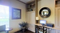 This single-floor space showcases a thoughtfully designed built-in workspace that serves as a functional and visually appealing focal point. The built-in unit features a combination of open shelving and cabinet storage with raised-panel doors, providing ample organization for books, decorative objects, and everyday essentials. The shelves display tasteful accents, including ceramic vases and stacked boxes, adding character to the space.

The integrated desk area offers a practical workspace, complete with a desktop computer setup, a classic table lamp with a warm cream shade, and a wooden chair with upholstered seating for comfort. Dark hardwood flooring grounds the room and contrasts elegantly with the soft, warm wall tones and crisp white cabinetry, enhanced further by detailed crown molding along the upper edges.

A large framed artwork on the wall adds a touch of sophistication, while a bold, round convex mirror with a dark frame introduces visual depth and a contemporary accent. A potted spiky plant near the base of the cabinetry brings a natural, organic element to the overall composition. The space conveys a refined, professional ambiance with a warm, welcoming character.