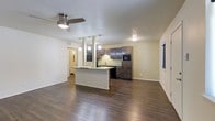 This single-floor space showcases an open-concept layout that seamlessly connects the living area and kitchen, creating a functional and cohesive environment. The kitchen is a standout feature, equipped with rich dark wood cabinetry that extends to the upper walls, providing ample storage. A built-in microwave and black appliances complement the cabinetry, while a dual-basin sink is positioned beneath under-cabinet lighting that illuminates the workspace effectively. A white peninsula island anchors the kitchen, offering additional counter space and serving as a natural divider between the cooking and living areas. Pendant lighting hangs above the peninsula, adding both task lighting and a decorative element. The living area is generously proportioned, featuring dark hardwood-style flooring that runs throughout the entire space, lending warmth and visual continuity. A ceiling fan with an integrated light fixture is centrally mounted, ensuring comfortable airflow. The textured ceiling and neutral, light-colored walls provide a clean backdrop. A window on the far wall introduces natural light into the living area, while a hallway visible in the background suggests access to additional rooms beyond.