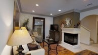 This three-floor space offers a thoughtfully designed interior that balances classic elegance with comfortable living. The living area showcased in the image is a welcoming room anchored by a striking white-mantled fireplace, which serves as the focal point of the space. Recessed lighting is embedded throughout the smooth ceiling, which is framed by crown molding, lending a refined architectural detail to the room.

The flooring consists of rich hardwood planks that add warmth and character to the overall aesthetic. Neutral-toned walls provide a versatile backdrop, with one accent wall in a deeper taupe shade adding visual depth. A large floor-length mirror with a dark wood frame reflects the room, enhancing the sense of openness. Décor elements include a sunburst mirror above the fireplace, an ornate dark wood media console, and lush indoor greenery that softens the space.

Seating arrangements include plush upholstered furniture styled with textured throws and accent pillows, centered around a glass-top coffee table. The overall ambiance is cozy yet polished, suggesting a well-curated interior spread across three functional floors.