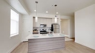 This contemporary single-floor residence is designed with a clean, modern aesthetic throughout. The open-concept kitchen serves as the heart of the home, showcasing white upper cabinetry paired with wood-toned lower cabinets, a subtle tile backsplash, and a full suite of stainless steel appliances including a range, microwave, and refrigerator. A large central island with a white quartz countertop and integrated sink provides ample prep space and casual seating, illuminated by elegant pendant lighting overhead. Light hardwood-style flooring flows through the main living areas, complementing the bright, neutral color palette. The generous living area is filled with natural light from multiple windows and offers direct access to a private balcony with open views of the surrounding landscape. The bedrooms are carpeted for comfort, each featuring windows that frame expansive outdoor views. A spacious walk-in closet provides well-organized storage with built-in shelving and hanging rods on both sides. Two full bathrooms are finished with wood-toned vanities, white quartz countertops, large mirrors, and shower-tub combinations. A dedicated laundry area houses a full-size side-by-side washer and dryer, adding everyday convenience to this well-appointed residence.
