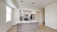 This contemporary single-floor residence is designed with a clean, modern aesthetic throughout. The open-concept kitchen serves as the heart of the home, showcasing white upper cabinetry paired with wood-toned lower cabinets, a subtle tile backsplash, and a full suite of stainless steel appliances including a range, microwave, and refrigerator. A large central island with a white quartz countertop and integrated sink provides ample prep space and casual seating, illuminated by elegant pendant lighting overhead. Light hardwood-style flooring flows through the main living areas, complementing the bright, neutral color palette. The generous living area is filled with natural light from multiple windows and offers direct access to a private balcony with open views of the surrounding landscape. The bedrooms are carpeted for comfort, each featuring windows that frame expansive outdoor views. A spacious walk-in closet provides well-organized storage with built-in shelving and hanging rods on both sides. Two full bathrooms are finished with wood-toned vanities, white quartz countertops, large mirrors, and shower-tub combinations. A dedicated laundry area houses a full-size side-by-side washer and dryer, adding everyday convenience to this well-appointed residence.