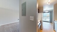 This single-floor unit offers a practical and straightforward layout with distinct living, sleeping, kitchen, and bathroom areas. The open living area is generously sized, finished with neutral carpet underfoot and anchored by a large sliding glass door that leads to a railed balcony, providing an outdoor view and abundant natural light. A wall-mounted heater unit is conveniently positioned within the living space.

The kitchen is efficiently arranged and features dark espresso-toned cabinetry with brushed hardware, light-toned laminate countertops, a coil-burner range with overhead ventilation, and a full-size refrigerator. Wood-style flooring runs through the kitchen, offering a warm contrast to the adjacent carpeted areas. A dishwasher is also present. An electrical panel and light switches are accessible near the kitchen entrance.

The bedroom is a well-proportioned, carpeted room with grey-painted walls and a wide horizontal window fitted with vertical blinds, allowing filtered natural light. A floor-level heating vent is built into the baseboard area.

The bathroom is clean and functional, featuring a white vanity with under-cabinet storage, a toilet, and a tub-shower combination with a curved curtain rod. Warm wood-tone flooring completes the space.