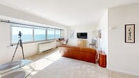 Located at 385 Ocean Blvd in Long Branch, NJ, this single-floor studio offers a well-organized layout that maximizes every square foot. The combined kitchen and living area forms the heart of the space, bathed in natural light from a wide bank of sliding windows that frame stunning, unobstructed views of the Atlantic Ocean — a defining feature that brings an airy, coastal ambiance throughout.

The living area features neutral carpet flooring, crisp white walls, and ample room for comfortable seating and a workspace positioned to take full advantage of the oceanfront panorama. The kitchen is thoughtfully laid out in a U-shape with white cabinetry extending to the ceiling, light countertops, a gas range with oven, a full-size refrigerator, and a peninsula that creates a natural division between the kitchen and living space. Warm tile flooring grounds the kitchen zone. A dedicated closet provides additional storage, enhanced by a striking navy blue armoire that adds a bold design accent. The bathroom is clean and functional, finished in white with mosaic tile flooring, a vanity with under-sink storage, and a mirrored medicine cabinet. Overall, the space exudes a simple, bright coastal character suited to relaxed seaside living.
