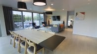 This single-floor space showcases a thoughtfully designed open-concept layout that fluidly connects the dining and living areas. The dining area is anchored by a substantial two-tone table — featuring a light wood surface paired with a dark contrast insert — surrounded by matching light wood chairs with dark upholstered seats. Overhead, bold cylindrical pendant lights in dark finishes add a striking visual element to the space.

The living area beyond is furnished with a coordinated arrangement of navy blue accent chairs, a grey sofa accented with an orange throw, and a geometric-patterned area rug. A wall-mounted flat-screen television serves as a focal point. Floor lamps provide ambient lighting throughout the seating area, while recessed ceiling lights illuminate the space uniformly.

Light hardwood-style flooring runs continuously throughout, enhancing the sense of openness. Large floor-to-ceiling sliding glass doors along one wall flood the interior with natural light and provide direct views and access to an outdoor patio area. The neutral grey-toned walls complement the contemporary furnishings, creating a cohesive and modern aesthetic throughout.
