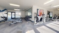 Located in Tustin, CA, this fitness center offers a generously sized, single-floor exercise room designed to support a wide range of workout routines. The space features a striking herringbone-patterned carpet throughout, lending a modern aesthetic that complements the clean, white walls and bright, flush-mounted lighting. Large windows along multiple walls invite ample natural light and provide views of the surrounding outdoor environment, creating an energizing atmosphere for exercise.

The facility is thoughtfully organized into distinct workout zones. A strength training area includes a full rack of dumbbells displayed on tiered stands, accompanied by a large wall mirror that enhances the sense of space. Multiple HOIST-brand cable and resistance machines are positioned throughout, alongside a weight bench and leg press equipment. A dedicated functional training zone houses a BOX-branded power rack with suspension trainers, a heavy punching bag, and battle ropes, catering to dynamic and combat-style workouts. A ceiling fan and exposed HVAC ductwork add industrial character to a separate training section.

Colorful abstract artwork decorates the walls, adding personality and visual interest. The open floor plan, high vaulted ceilings in select areas, and well-spaced equipment arrangement ensure comfortable movement throughout this comprehensive fitness facility.
