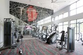 Located on Wood Street SE in Atlanta, Georgia, this well-appointed fitness center offers a spacious, single-floor layout with impressive double-height ceilings that create an open, energizing atmosphere. A dramatic wall of floor-to-ceiling windows lines one side of the space, flooding the interior with natural light and providing views of the tree-lined streetscape outside. The design is further elevated by a bold graphic mural — a stylized city map rendered in red and black — that commands attention on the upper wall, reinforcing a distinct urban identity. Underfoot, patterned carpet tiles in grey with bold red accent stripes add a dynamic visual element throughout the space. A striking black spiral staircase connects the main floor to an upper-level mezzanine, which overlooks the gym below through glass railings, adding architectural depth and dimension. The equipment offering is comprehensive, including a full row of treadmills, ellipticals, and other cardio machines positioned along the window wall, complemented by an extensive selection of strength-training machines, cable systems, free weights, and adjustable benches. Mounted flat-screen televisions are integrated throughout for entertainment during workouts. The overall ambiance is modern, functional, and motivating — a well-designed facility suited for a complete fitness experience.