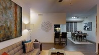 This single-floor residence showcases an open-concept design that seamlessly connects the living, dining, and kitchen areas into one cohesive, flowing space. The living area is anchored by a comfortable upholstered sofa accented with teal and gold throw pillows, complemented by a classic table lamp and a geometric wire wall sculpture that serves as a notable artistic focal point. Warm-toned hardwood flooring runs throughout, adding continuity and a sense of warmth to the space. A ceiling fan overhead provides both function and subtle style. The kitchen is partially open to the main living area, visible through a pass-through opening, and features a breakfast bar with tall dark bar stools, under-cabinet lighting, and what appears to be dark cabinetry. Adjacent to the kitchen, a dedicated dining area is furnished with a dark wood table and matching chairs, illuminated by track lighting mounted to the ceiling. Decorative panel artwork lines the dining area wall, adding texture and visual interest. The overall ambiance is modern and curated, balancing contemporary décor with warm, inviting tones that create a comfortable and stylish living environment.