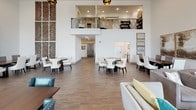 Located at 3850 Sycamore School Rd in Fort Worth, Texas, this single-story community space is designed with an open, airy layout that prioritizes comfort and social interaction. The interior is characterized by dramatically high ceilings that amplify the sense of scale throughout, with crisp white walls providing a clean, modern backdrop.

The main gathering area is expansive, accommodating multiple distinct zones including a dining area furnished with round tables and upholstered chairs, and a comfortable lounge section featuring plush sofas, accent chairs, and a wall-mounted flat-screen television. Wood-look tile flooring runs throughout, lending warmth and continuity to the space.

Natural light pours in through oversized, floor-to-ceiling windows framed with elegant drapery, offering views of the surrounding landscaped exterior. A mezzanine level with decorative iron railings is visible from the main floor, adding architectural interest and vertical depth to the room.

The kitchen area is outfitted with dark cabinetry, a subway tile backsplash, built-in appliances, and a generously sized island with seating — ideal for casual gatherings. Decorative pendant lighting and textured wall panels add refined finishing touches, contributing to an overall ambiance that is both welcoming and polished.