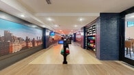 Located at 5180 Peachtree Blvd in Chamblee, Georgia, this dedicated bowling alley delivers a polished and energetic entertainment experience within a well-appointed single-floor layout. The space features two full-length bowling lanes finished in smooth light hardwood, flanked by dark gutters and equipped with modern ball-return stations stocked with a colorful selection of bowling balls. Prominent wall murals depicting dramatic urban skylines line the lanes, creating an immersive, city-inspired atmosphere, while bold "Great Gatsby"-themed neon signage adds a retro, theatrical flair. Dark navy-painted brick columns and accent walls provide architectural contrast against the light flooring and white ceiling outfitted with recessed lighting throughout. A dedicated ball and shoe rack station is built into the sidewall, offering organized storage for equipment. At the far end of the lanes, a comfortable lounge area features a large wraparound white upholstered bench, curated artwork displays, and a dark countertop bar with leather bar-stool seating — ideal for spectators and socializing. The seamless flow between the active lane area and the relaxed lounge zone makes this space well-suited for both casual recreation and private events.