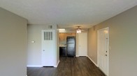 This single-floor space features an open-concept layout that seamlessly connects the living area to the kitchen, creating an efficient and functional floor plan. Dark hardwood-style flooring extends throughout, providing a rich contrast against the neutral beige walls and bright white trim and baseboards.

The living area is straightforward and versatile, offering ample room for furnishing. A ceiling fan with a light fixture is centrally positioned, contributing both airflow and ambient lighting. A louvered closet door on one wall provides convenient storage, while a standard entry door with a paneled design is situated nearby, offering direct access to the unit.

The kitchen, visible from the main living space, is equipped with black appliances including a full-size refrigerator and dishwasher. Light wood cabinetry lines the upper walls, providing storage, and under-cabinet lighting adds functional illumination to the workspace. A wall-mounted HVAC vent is positioned above, ensuring climate control throughout the space. The textured ceiling adds a subtle visual detail consistent throughout the unit.