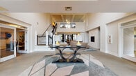 Located at 2110 Cinema Dr in Fuquay-Varina, NC, this contemporary space makes an immediate impression with its dramatic double-height ceilings and expansive, open-concept layout. The main living area is anchored by a sleek built-in fireplace framed by symmetrical display shelving with teal-accented insets, complemented by curved modern sofas and a glass-top coffee table resting on a large geometric area rug. A striking staircase with black metal railings serves as a central architectural feature, connecting the main floor to an upper mezzanine level and adding vertical drama to the space. Pendant globe lighting and coffered ceiling details contribute to the refined yet contemporary atmosphere. The kitchen area is well-appointed with white cabinetry, granite countertops, a large island with bar seating in bold orange upholstery, pendant lighting, and stainless steel appliances—opening directly to a casual dining nook. A separate lounge area offers an energetic contrast, featuring an eye-catching orange accent wall adorned with vibrant pop-art panels, exposed industrial-style ductwork overhead, and an assortment of modern accent seating. The overall design blends modern architecture with eclectic, colorful interior styling to create a dynamic and functional communal environment.