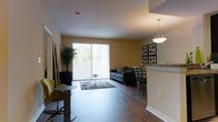 This single-floor residence showcases a thoughtfully designed open-concept layout that integrates the living, dining, and kitchen areas into one cohesive, flowing space. Rich hardwood flooring runs throughout, adding warmth and a sense of continuity to the interior. The living area is anchored by a large sliding glass door fitted with vertical blinds, flooding the space with natural light and providing access to an outdoor area beyond. A plush area rug and comfortable seating create a relaxed, inviting atmosphere in the lounge zone. Adjacent to the living area, the dining space accommodates a table and chairs, illuminated by a stylish pendant light that adds a decorative focal point overhead. A partial wall with a granite-topped counter defines the kitchen boundary, offering a semi-open pass-through feel while maintaining a sense of separation. Warm beige-toned walls complement the wood floors and neutral furnishings throughout. Framed wall art adds a personalized touch to the dining area. The space benefits from clean lines, smooth ceilings with recessed lighting, and an efficient single-floor plan that makes the interior feel spacious and practical for everyday living.