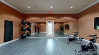 Located at 423 Highway 466 in Lady Lake, Florida, this dedicated exercise room offers a focused and motivating environment for fitness activity. The space is finished with warm terracotta-toned walls that create an energizing atmosphere, complemented by clean white crown molding and baseboards throughout. Recessed lighting illuminates the room evenly, highlighting the smooth, light-toned laminate flooring that extends across the entire area.

A full-length wall mirror spans one side of the room, enhancing the sense of space and providing visual feedback during workouts. A large flat-screen television is mounted on the wall, ideal for following along with guided exercise sessions. The room is equipped with multiple stationary spin bikes arranged for group cycling classes, along with a rack of medicine balls, stability balls on a dedicated stand, and wall-mounted exercise mats for floor-based training.

A motivational quote — "Persistence can change failure into Extraordinary Achievement" — is prominently displayed in elegant script along the upper wall, adding an inspirational element to the space. A glass-paneled door provides access and allows natural light to filter in. The room is designed to accommodate a variety of fitness routines, making it a versatile and well-appointed facility for residents and users in the Lady Lake community.