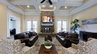 The clubhouse lounge is a thoughtfully designed communal space that blends classic architectural detail with comfortable, inviting furnishings. The room's most striking feature is a floor-to-ceiling stacked stone fireplace, which serves as the focal point of the space and adds a warm, rustic elegance to the overall aesthetic. A large flat-screen television is mounted above the fireplace mantel, making the area well-suited for both casual relaxation and group viewing.

The ceiling draws immediate attention with its elegant coffered design, accented by recessed lighting and a ceiling fan that contributes to both the visual interest and comfort of the room. French doors flank either side of the fireplace wall, inviting natural light into the space and providing views of the surrounding exterior.

The seating arrangement centers around rich, dark leather sofas paired with patterned accent chairs, all anchored by a wood coffee table and a layered area rug. Potted greenery is thoughtfully placed throughout, adding a fresh, natural touch. The warm neutral tones of the walls, combined with the wood flooring and stone accents, create a cohesive and sophisticated ambiance ideal for community gatherings.