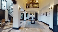 The Cortland Clubhouse in Boca Raton, FL, is an elegantly designed two-story amenity space that blends Mediterranean-inspired architecture with modern, upscale interiors. The grand entrance hall sets the tone immediately, featuring soaring vaulted ceilings, polished travertine flooring with inlaid wood detailing, and an ornate wrought iron chandelier suspended above a central round table. A sweeping staircase with intricate ironwork balustrades leads to the upper level, which overlooks the foyer through a decorative wrought iron balcony railing.

The main level offers multiple gathering areas, including a sophisticated lounge with a dramatic double-height fireplace, arched windows with wrought iron accents, and refined seating arrangements. A separate social lounge opens to the outdoors through French doors, offering natural light and comfortable furnishings. The fully equipped demonstration kitchen features rich dark cabinetry, quartz countertops, a farmhouse sink, patterned tile backsplash, and integrated televisions. A dedicated screening room with coffered ceilings, projection capabilities, and plush oversized seating rounds out the amenities, delivering a refined, resort-style clubhouse experience.