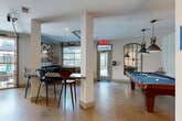 Located at 747 Ralph McGill Blvd NE in Atlanta, GA, this single-floor amenity lounge is a well-appointed communal space that blends modern design with a lively, social atmosphere. The open-concept layout is anchored by polished concrete floors that span the entire area, lending an industrial-chic character throughout.

The lounge is divided into distinct functional zones. A dedicated billiards area features a full-size pool table illuminated by a pair of dramatic industrial pendant lights, complemented by bold artwork on the walls. Adjacent to this, a comfortable seating lounge showcases a striking three-dimensional textured white accent wall, a wall-mounted flat-screen TV, plush sofas arranged around a wood-and-metal coffee table, and a vibrant striped area rug. Mustard-yellow wingback chairs are positioned near large floor-to-ceiling windows that flood the space with natural light and offer outdoor views of the surrounding Atlanta streetscape.

A bar-height table with modern chairs provides an additional gathering spot, while a round marble-top dining table with sleek leather chairs adds a refined touch, accented by a Sputnik-style chandelier and industrial wall art. Large glass garage-style doors connect the interior to an outdoor patio area, enhancing the sense of openness and the flow between indoor and outdoor spaces.
